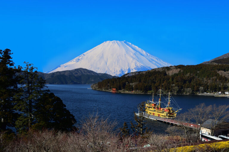 Gunung Fuji yang tertutup salju di atas Danau Ashi di Hakone, dengan kapal wisata di dermaga dan torii merah dari Kuil Hakone di tepi pantai