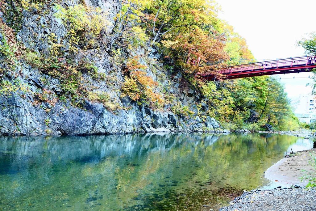 A red suspension bridge over the river at Jozankei, with the trees on the cliff turning yellow and orange A red suspension bridge over the river at Jozankei, with the trees on the cliff turning yellow and orange