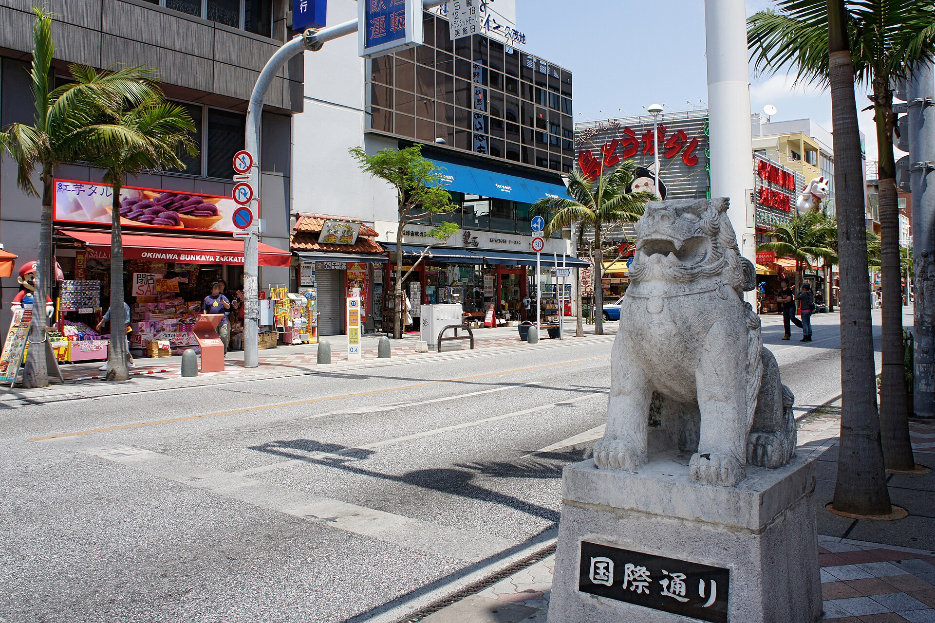 Kokusai-dori in Naha, Okinawa, with a shisa lion statue and street sign beside palm-lined shops
